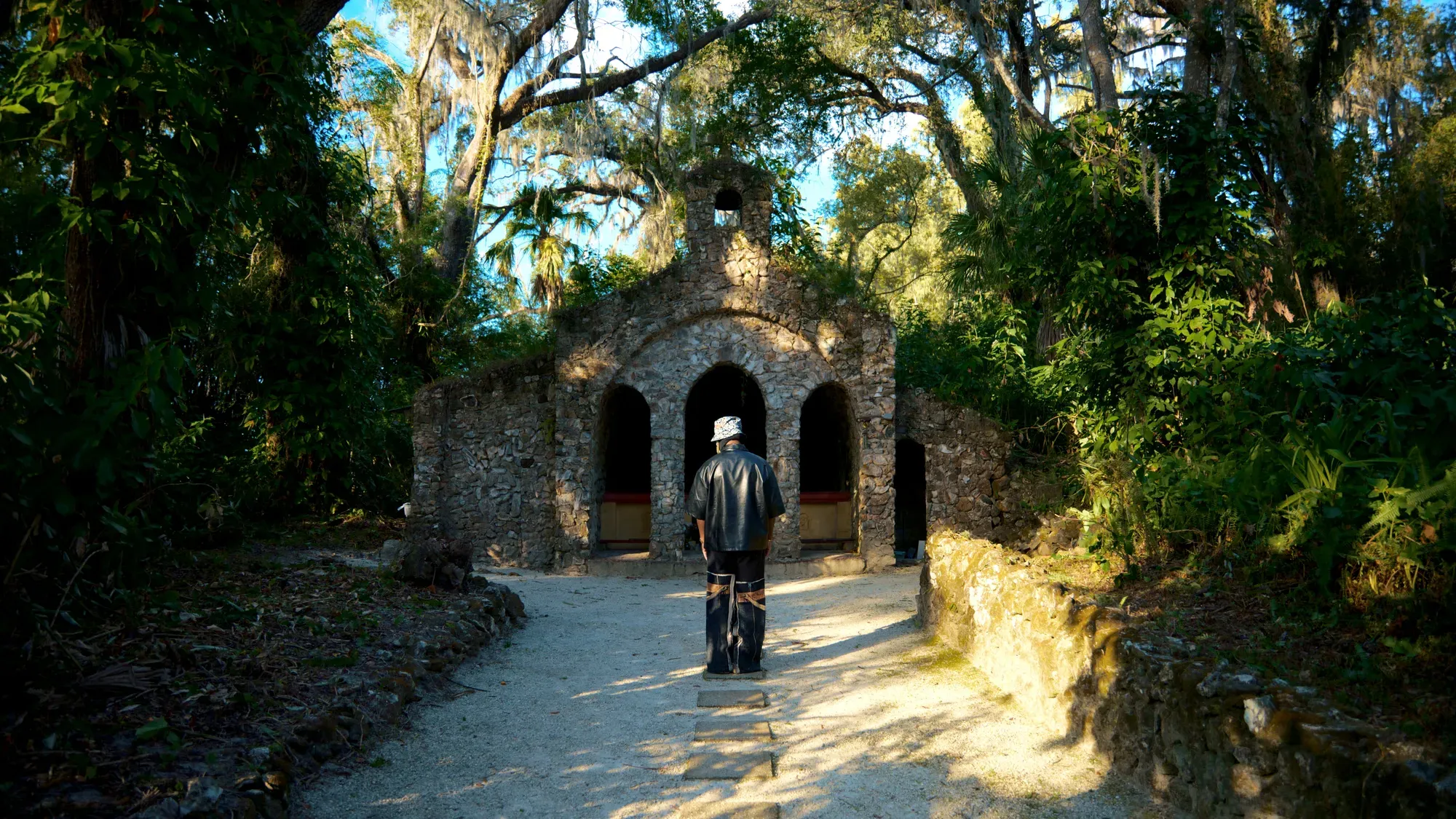 Pusha Preme standing near weathered stone architecture surrounded by greenery.