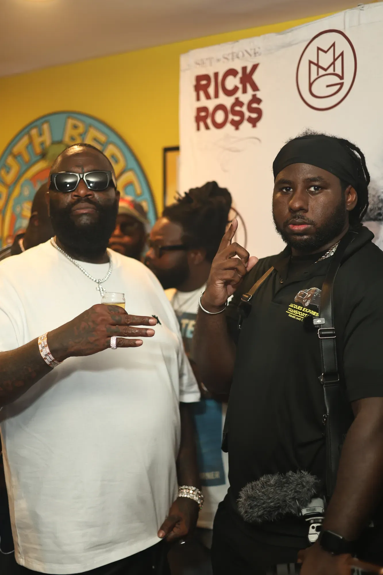 Rick Ross and another man in front of a Rick Ross and MMG backdrop.