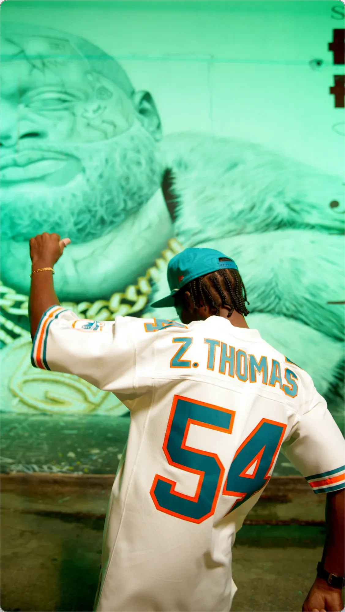 INIKIO from behind, the Z. Thomas #54 jersey toward the camera, in front of the green-lit mural.